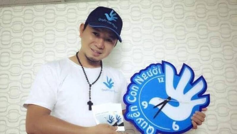 Das Foto zeigt Bùi Tuấn Lâm, der in seiner linken Hand eine große Uhr hält, auf die er mit seiner rechten Hand zeigt. Er trägt eine Baseballkappe und lächelt leicht in die Kamera.