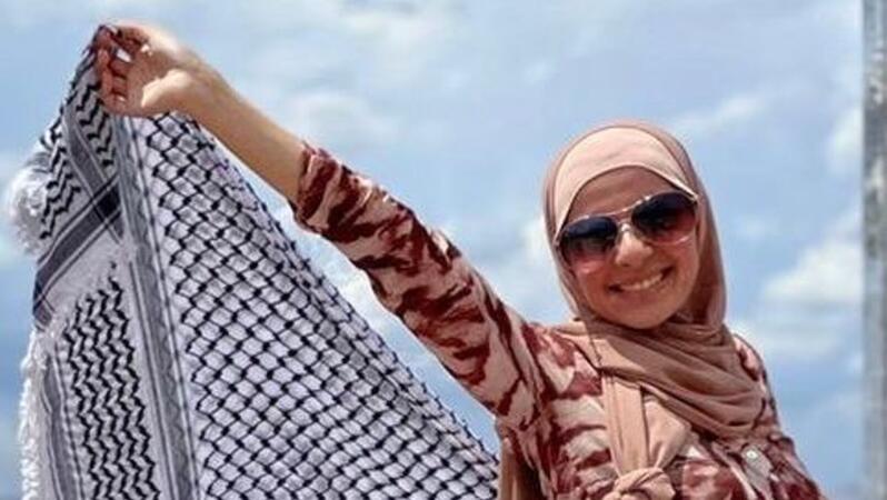 Das Foto zeigt Leqaa Kordia. Sie trägt ein Kopftuch und eine Sonnebrille und lächelt in die Kamera. Sie breitet mit ihren Händen hinter ihrem Rücken ein großes Kufiya-Tuch aus.