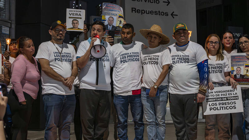 Eine Gruppe von Menschen bei einer Kundgebung, eine Person hält ein Megafon, sie tragen weiße T-Shirts mit der Aufschrift: "Befreit alle politischen Gefangenen"