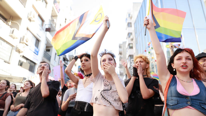 Das Foto zeigt mehrere Personen auf einer Kundgebung. Sie rufen Slogan und halten Regenbogenfahnen in der Hand. Einige lachen.
