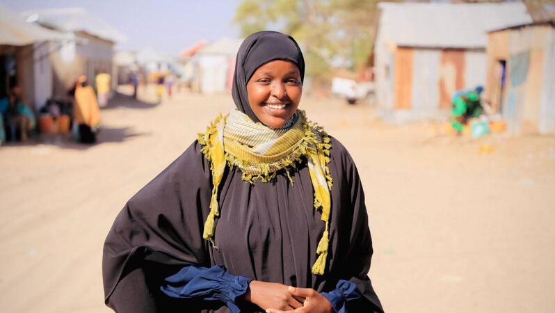 Das Foto zeigt Sadia Moalim Ali auf einer nicht asphaltierten Straße. Sie trägt ein Kopftuch und ein Gewand und lächelt in die Kamera. Im Hintergrund stehen einstöckige Gebäude.