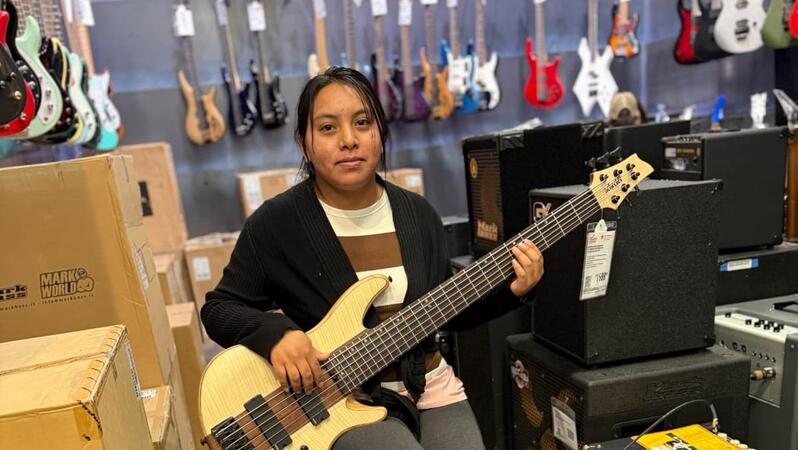 Das Foto zeigt Andrea Pedro-Francisco in einem Musikladen mit einem E-Bass in den Händen. Sie blickt in die Kamera und lächelt leicht. Hinter ihr hängen Gitarren an der Wand.